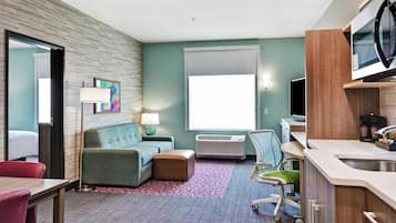 A hotel room with a sofa, a small table, a chair, a desk, a television, a window, and a kitchenette.