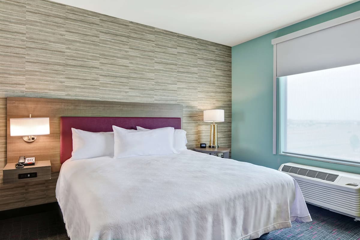 Une chambre d’hôtel avec un grand lit, une tête de lit en bois, une table de chevet avec une lampe, une autre table de chevet avec un téléphone et une fenêtre donnant sur un beau paysage.
