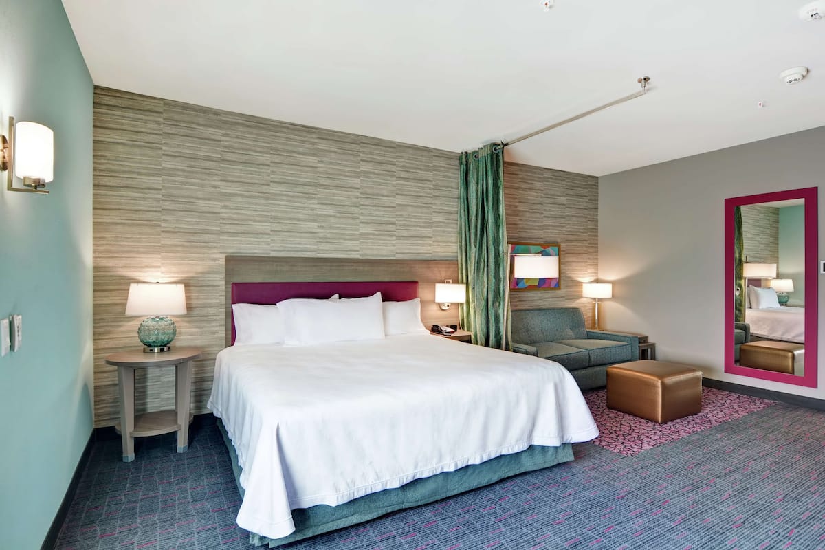 Une chambre d’hôtel avec un grand lit, deux tables de chevet avec des lampes, une chaise, un petit pouf et un miroir au mur.
