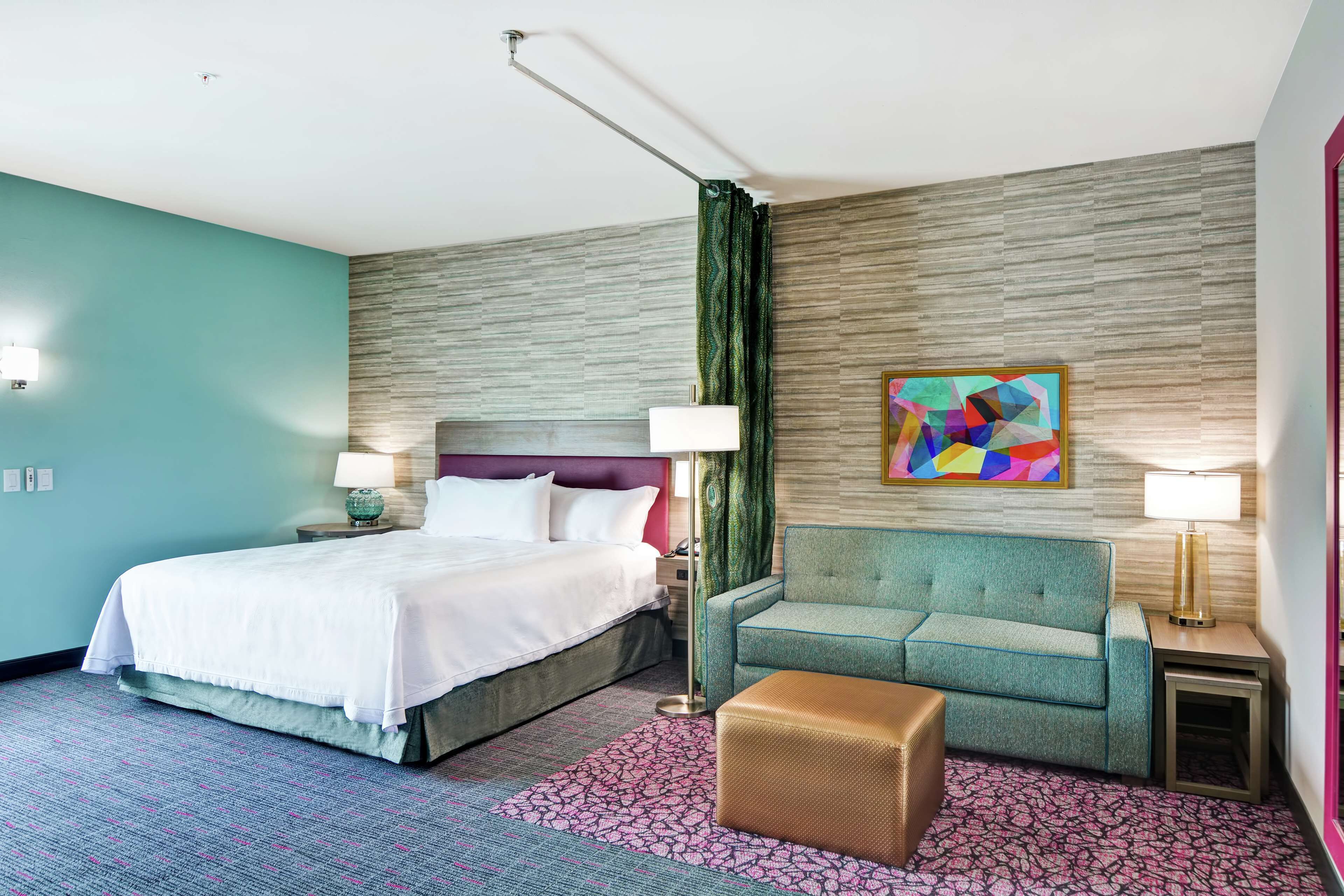 Una habitación de hotel con cama, sofá, una mesita, lámpara y un cuadro abstracto colorido en la pared.