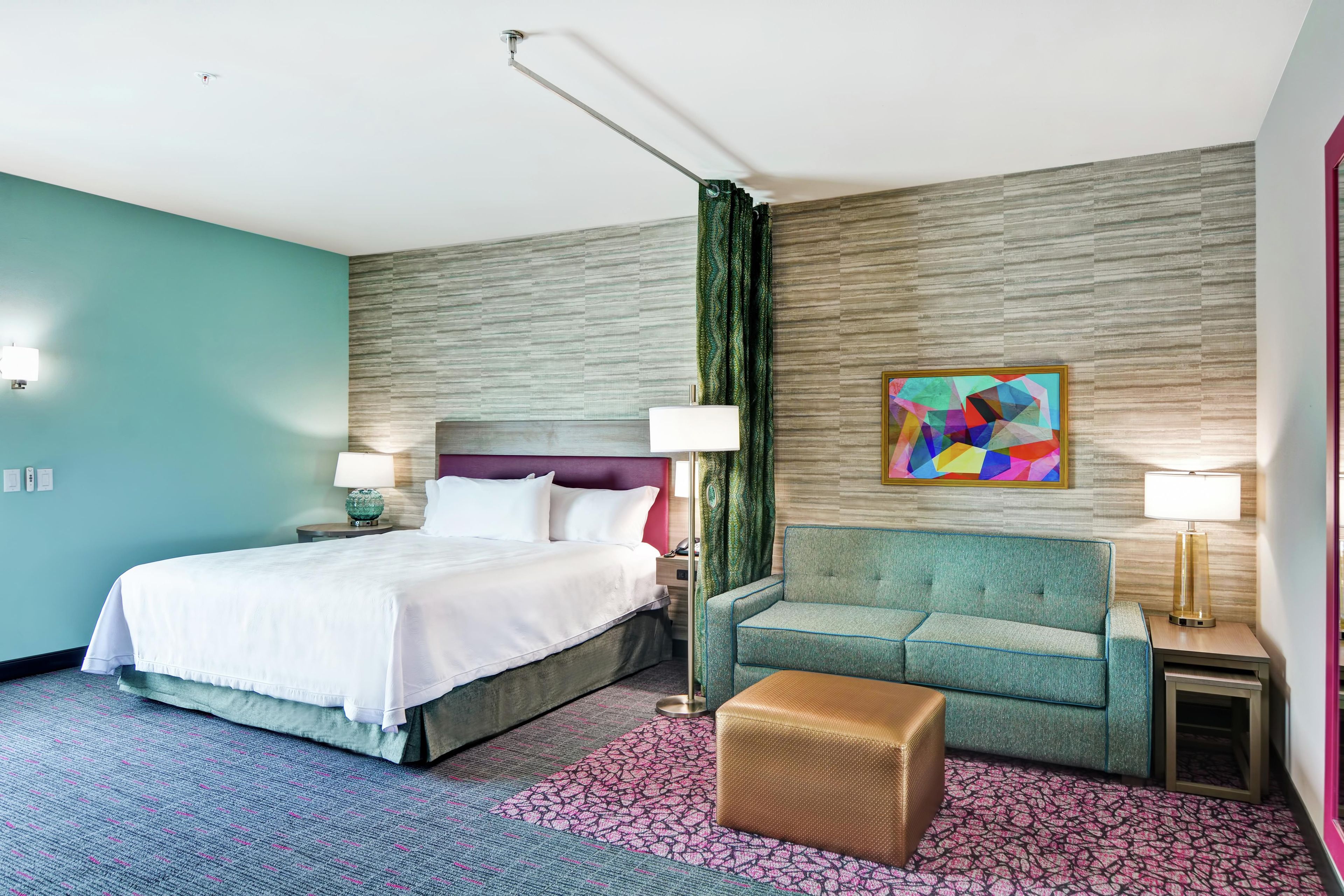 Una habitación de hotel con cama, sofá, reposapiés pequeño, un cuadro y una alfombra estampada.