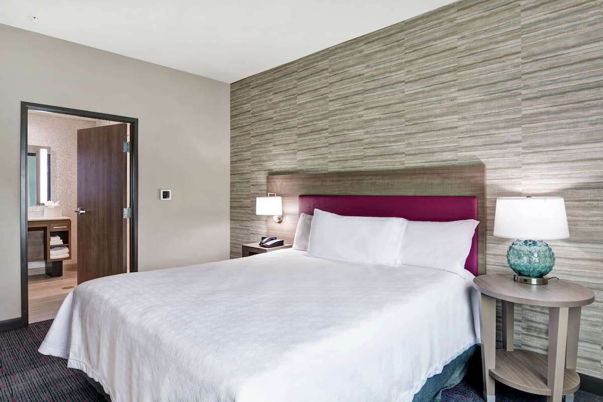 Une chambre d’hôtel avec un grand lit, une tête de lit en bois, une table de chevet avec une lampe et une porte donnant sur une salle de bain.