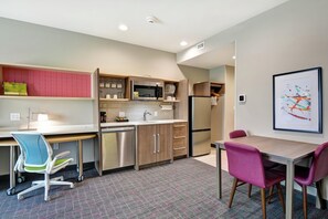 A compact kitchen with a microwave, refrigerator, and built-in oven. There is a dining table with chairs and a small office area with a desk and chair. - Home2 Suites by Hilton Pecos (Pecos)