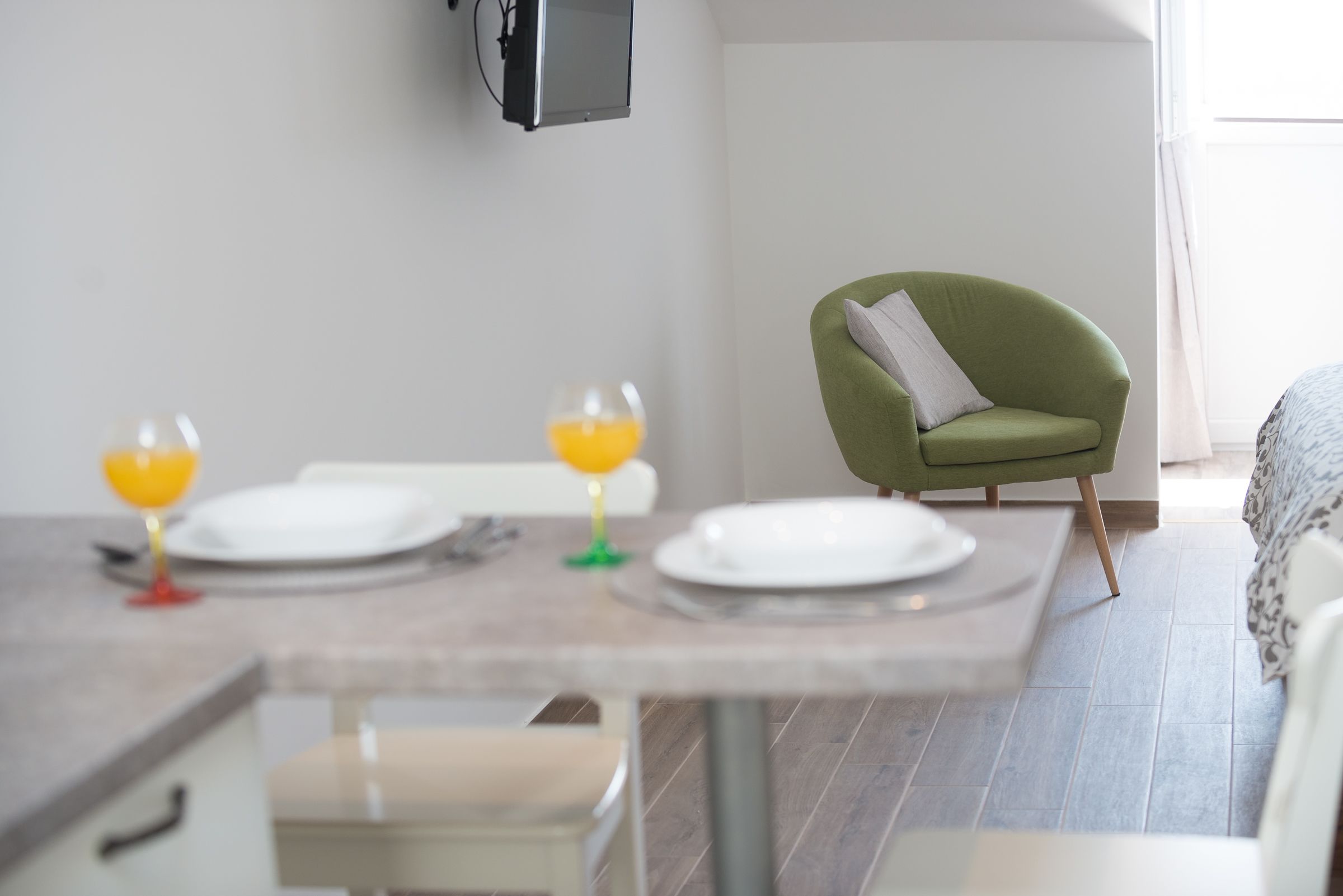 Un comedor con una mesa dispuesta para dos personas, dos vasos de jugo de naranja y un sillón verde con un cojín.