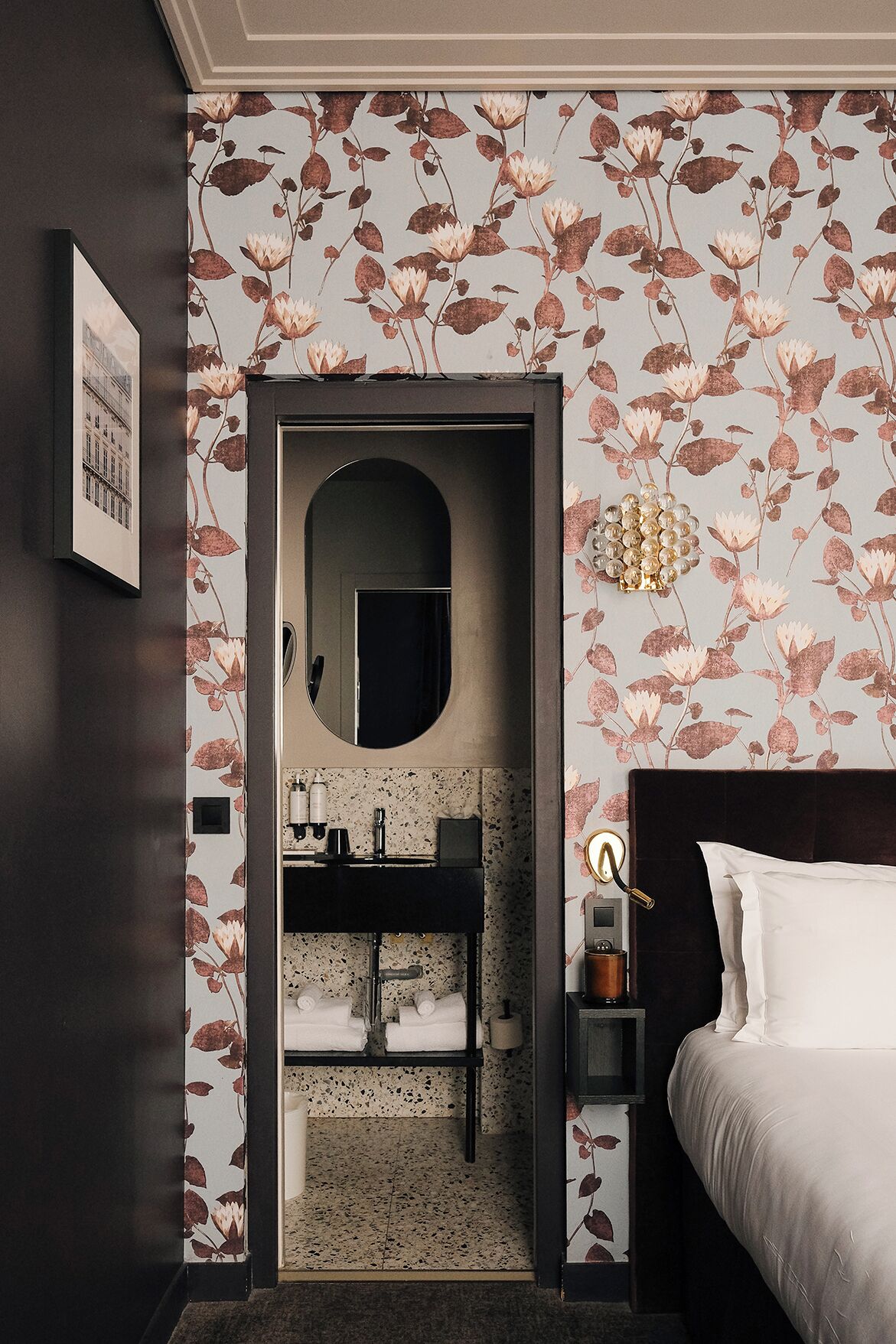 Un dormitorio con cabecera oscura, una cama, una mesita de noche, un espejo y un baño con lavabo y perchero.