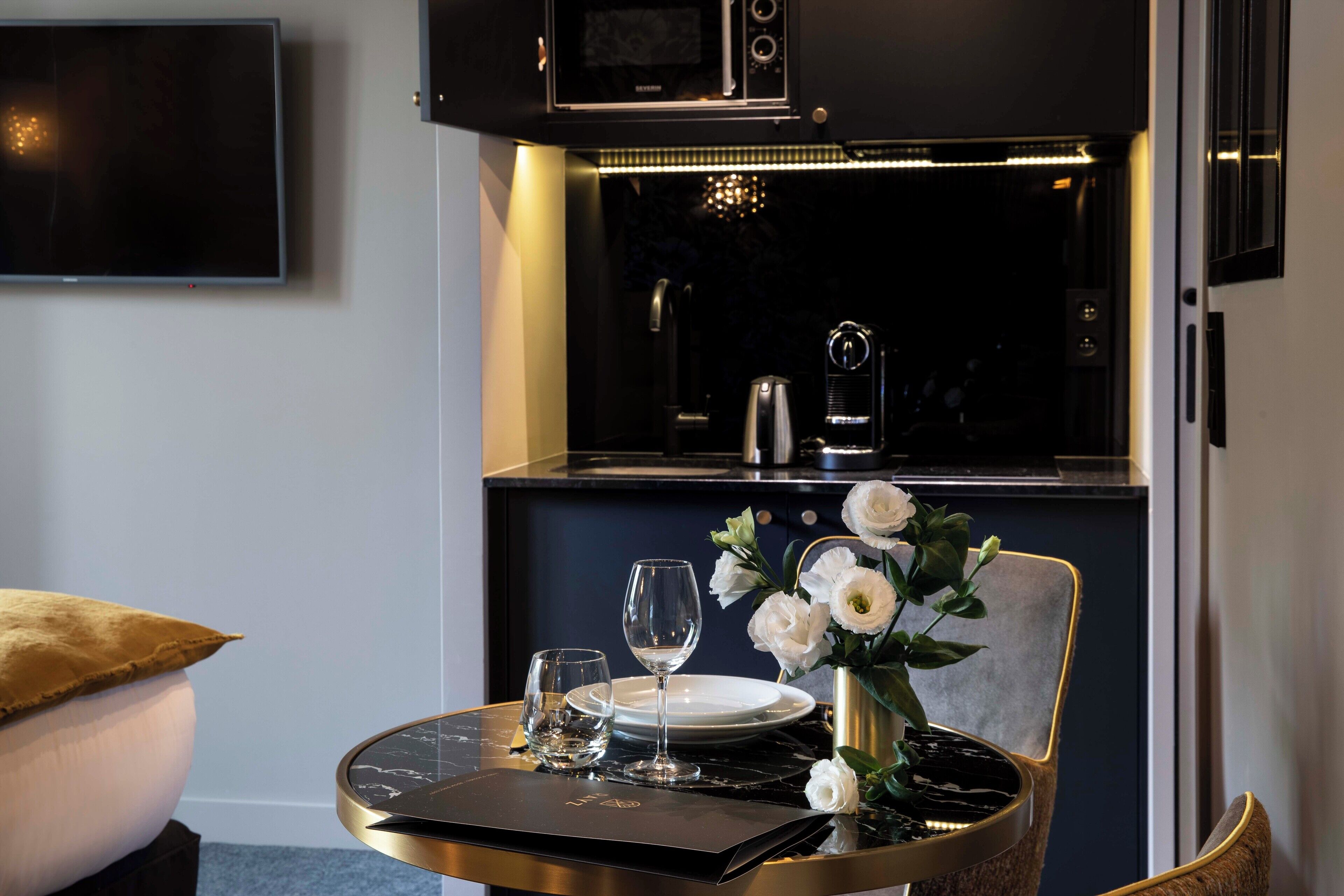 A dining area with a table set for two, a vase with white flowers, and a modern kitchen in the background.