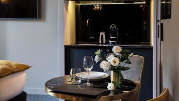 A dining area with a table set for two, a vase with white flowers, and a modern kitchen in the background.