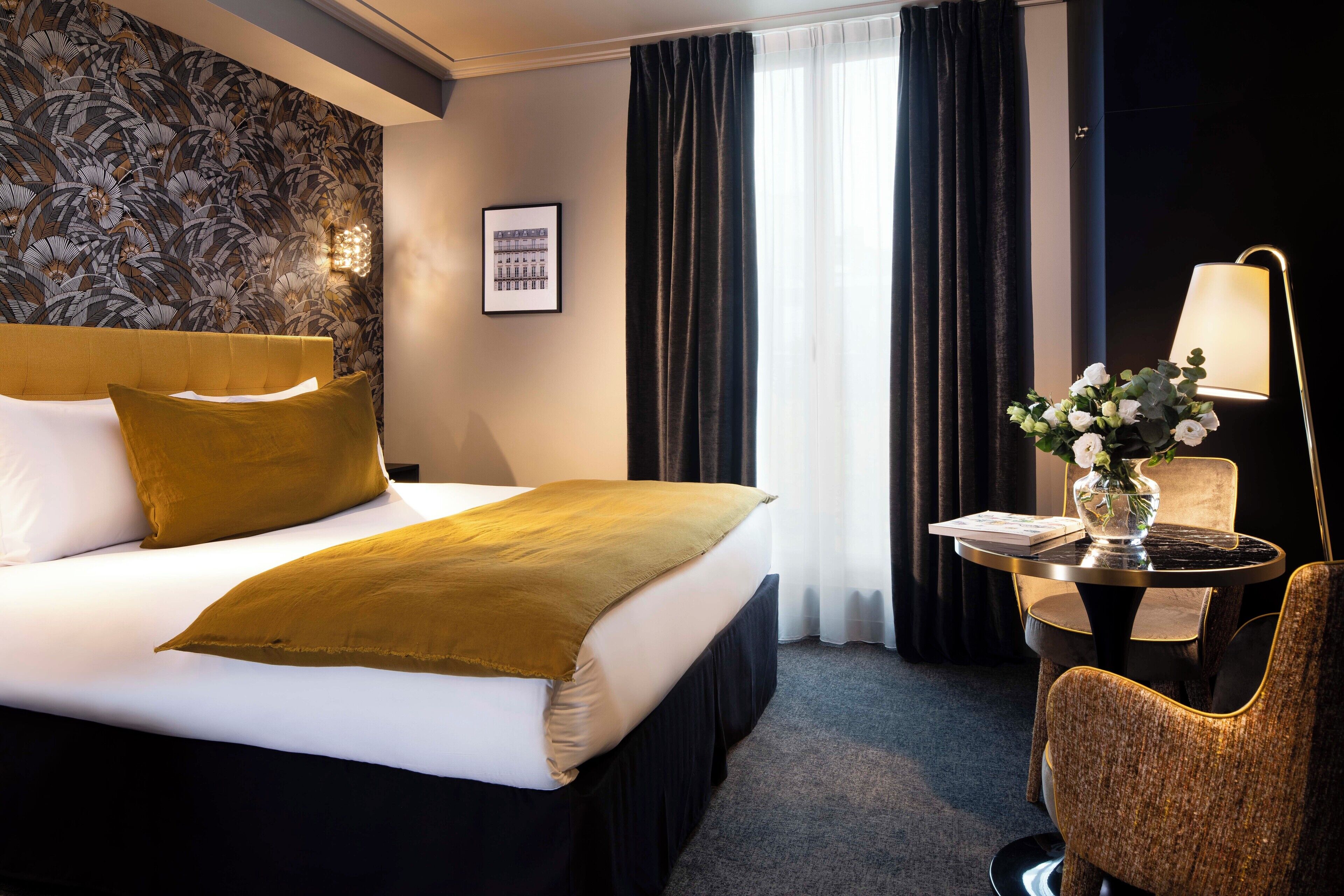 Ein Hotelzimmer mit einem großen Bett, einem kleinen runden Tisch mit Lampe, einem Sessel und einer Vase mit Blumen.
