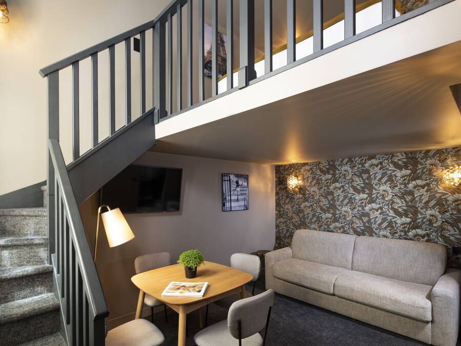 Un escalier menant à un niveau supérieur, un canapé, une table avec une plante et un mur recouvert d’un papier peint à motifs floraux.