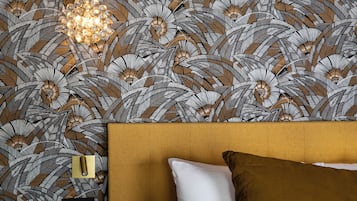 A room with a patterned wallpaper, a bed with two pillows, a nightstand with a lamp, and a power outlet.