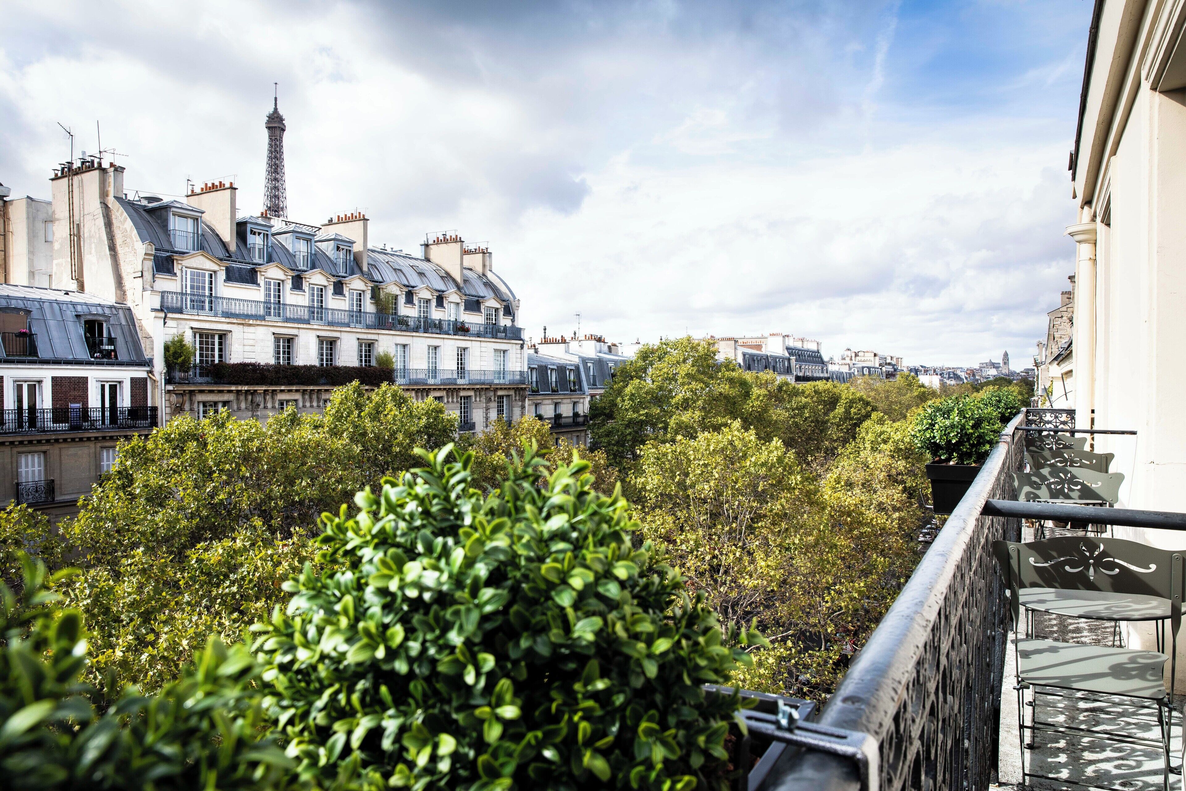 Une vue sur les bâtiments parisiens, avec la Tour Eiffel en arrière-plan, un balcon avec une balustrade et de luxuriants arbres verdoyants.