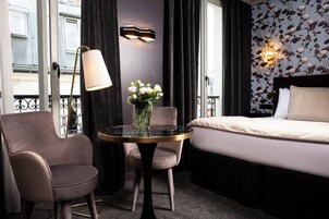 A hotel room with a bed, a round table with a vase of flowers, two chairs, a lamp, and a wall with floral wallpaper.