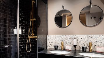 A modern bathroom with black tiled walls, a gold showerhead, two round mirrors, and a granite countertop with two sinks.