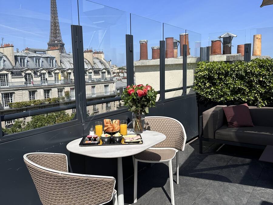 A rooftop terrace with a round table set for breakfast, wicker chairs, and a view of the Eiffel Tower.