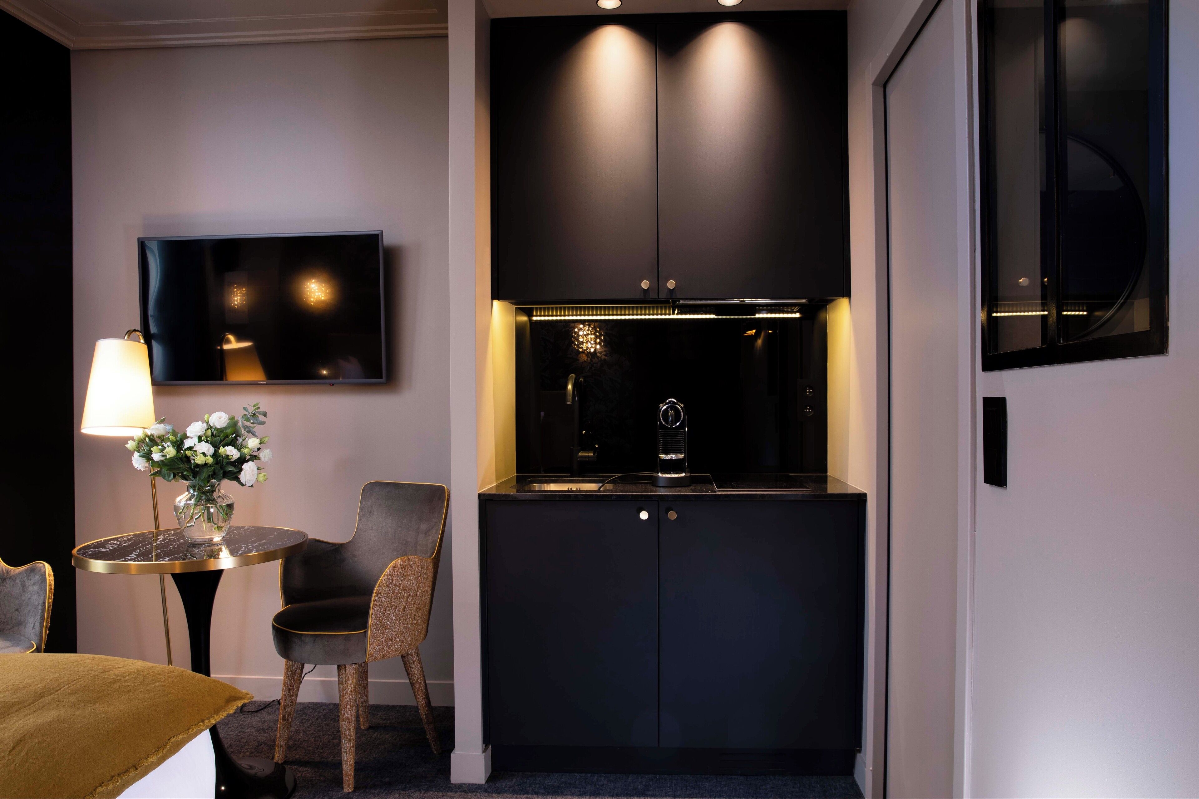 Habitación de hotel compacta con kitchenette, un televisor de pantalla plana montado en la pared, una zona de comedor con mesa y sillas, y una cama con cabecera color mostaza.