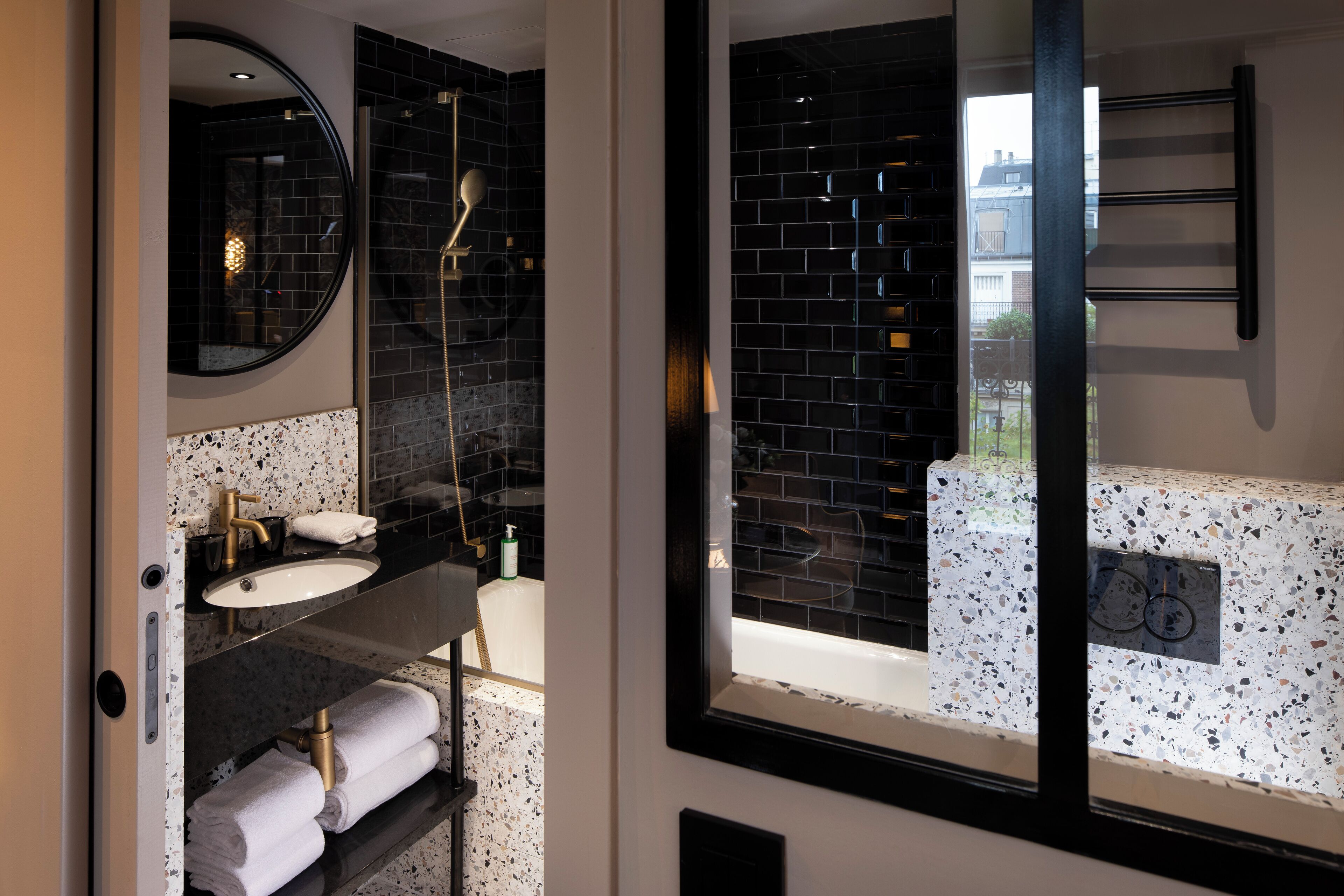 A modern bathroom with a round mirror, a sink with a gold faucet, and a bathtub visible through a window.