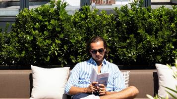 Un hombre sentado en un sofá marrón, leyendo un libro y con una taza sobre la mesa frente a él. Hay dos pequeñas macetas con plantas sobre la mesa.