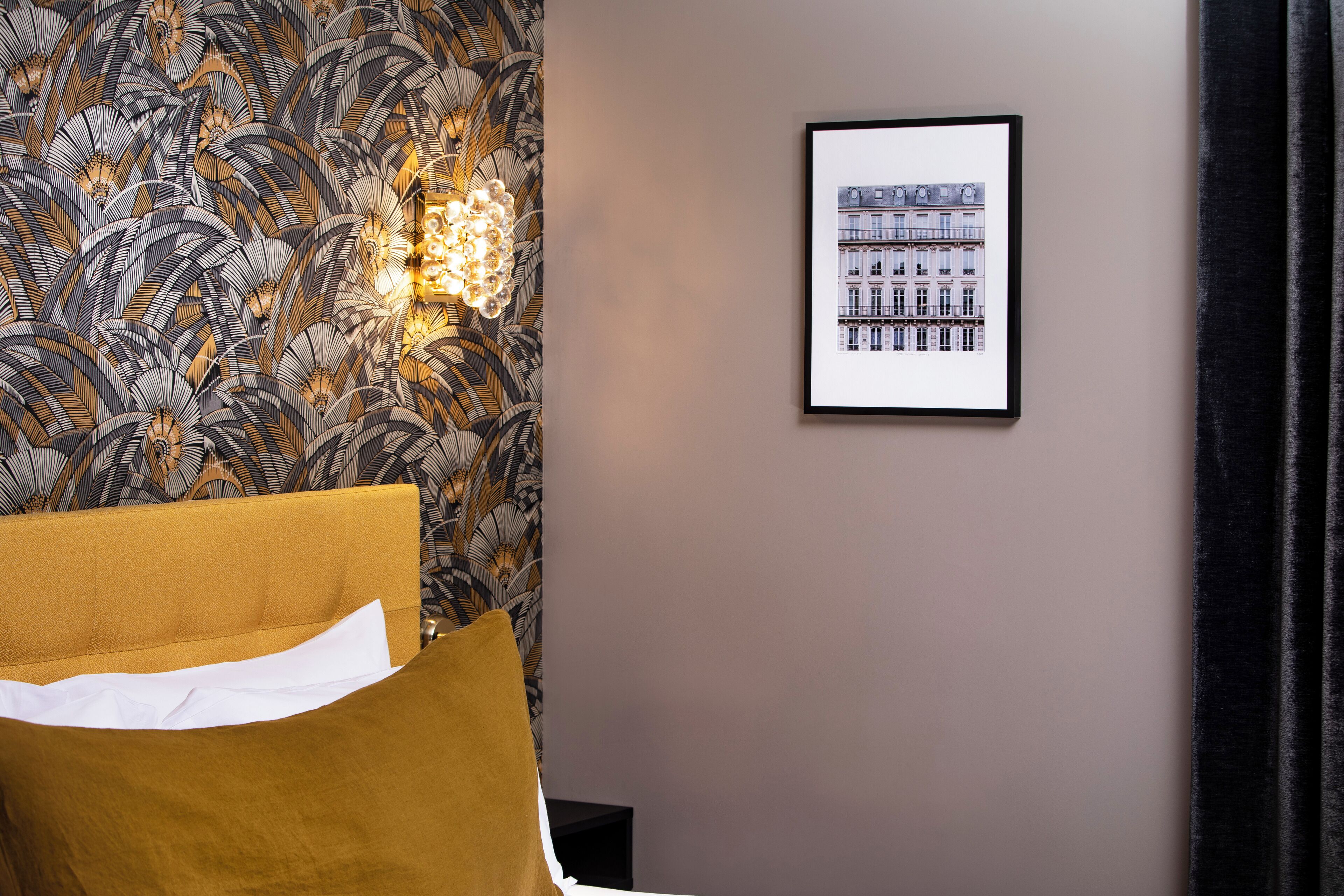 Habitación de hotel con papel tapiz estampado, un cuadro enmarcado de edificios, una cama con almohada blanca y una lámpara fijada a la pared.