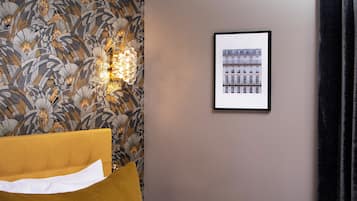 Habitación de hotel con papel tapiz estampado, un cuadro enmarcado de edificios, una cama con almohada blanca y una lámpara fijada a la pared.
