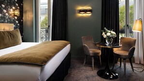 A hotel room with a bed, a table with chairs, a vase with flowers, and a wall-mounted light fixture.