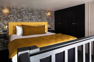 Un letto rifatto con cura, una coperta color senape, una testiera nera e una parete con motivo floreale.