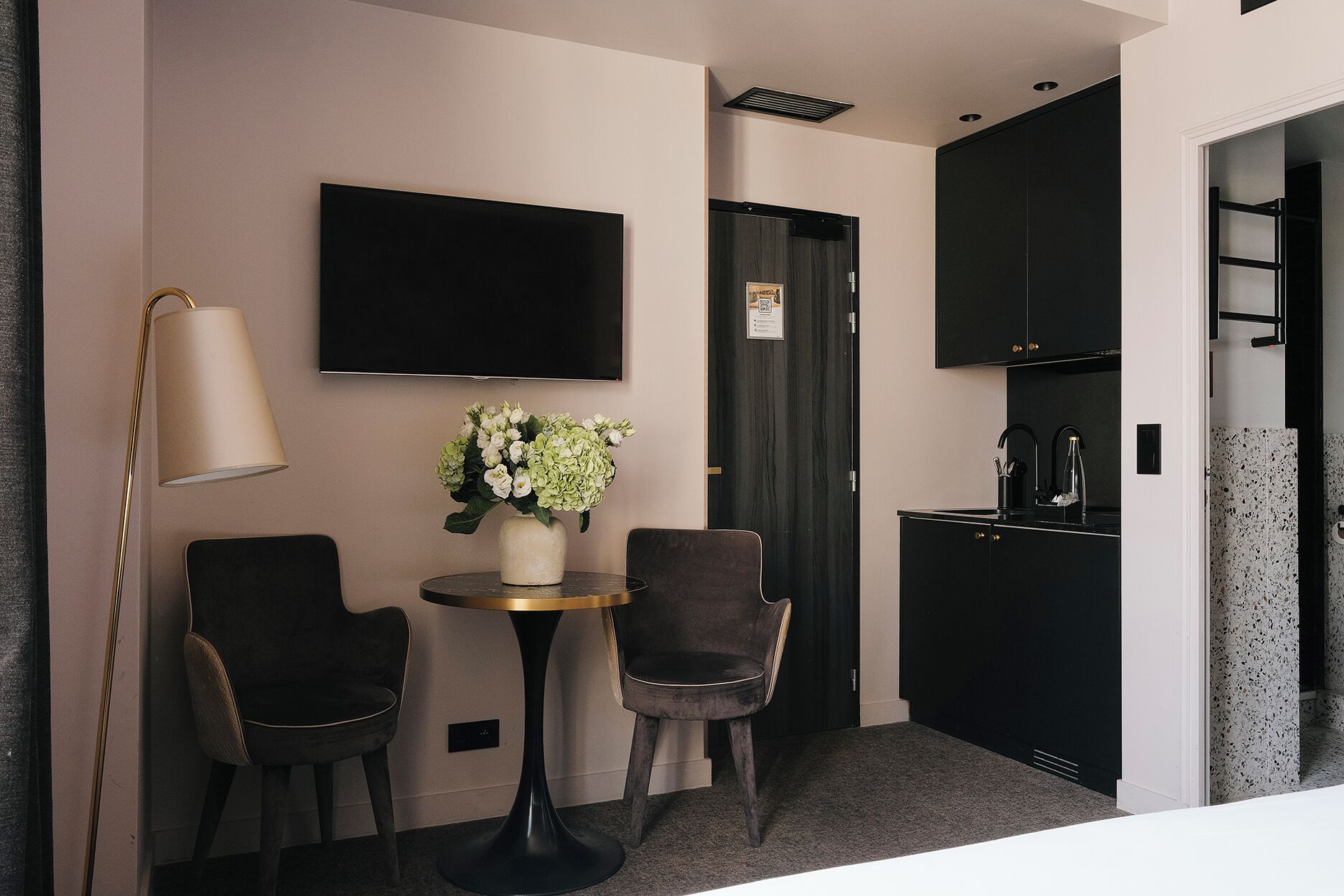 A compact hotel room with a flat-screen TV, a small round table with two chairs, a vase with flowers, a modern kitchen area, and a door with a sign.