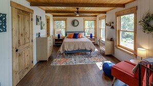 Un dormitorio con techo de madera, cama con almohadas anaranjadas y azules, una silla roja y una ventana con vista a árboles.
