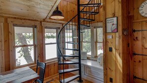 Interior de una cabaña de madera con una escalera de caracol, una mesa de comedor y vistas al exterior.