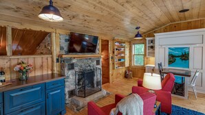 Interior acogedor de una cabaña con chimenea de piedra, paredes de madera, televisor montado encima y un sillón rojo.