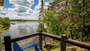 Una terraza de madera con una silla Adirondack azul que da a un lago con barcos, rodeado de árboles.