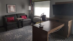 A modern hotel room with a grey sofa, a wooden desk, a large flat-screen TV, and a window with a view. - Hampton Inn & Suites Pittsburgh New Stanton (New Stanton)