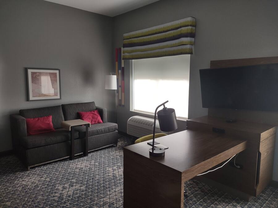 Habitació d'hotel moderna amb un sofà gris, un escriptori de fusta, un televisor pla gran i una finestra amb vistes.