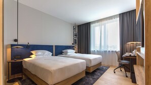 Ein Hotelzimmer mit zwei Betten, einem Schreibtisch, einem Sessel und einem großen Fenster mit Vorhängen.