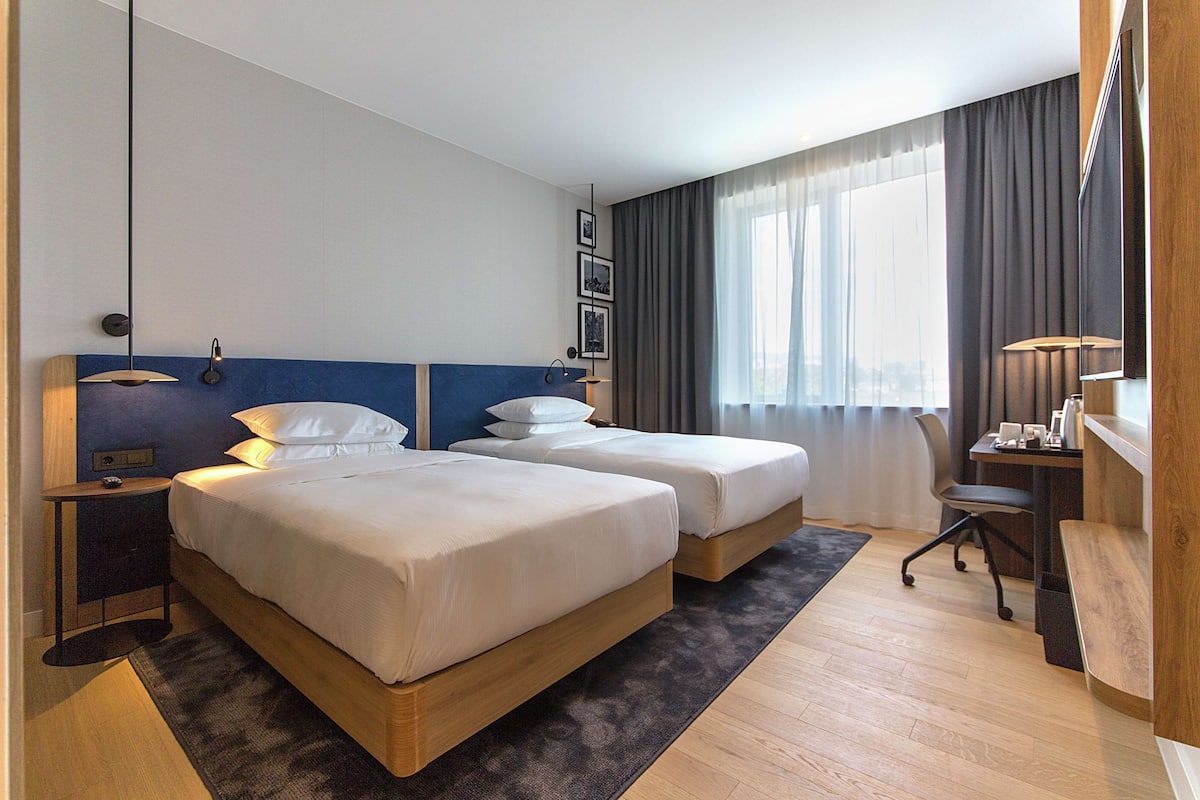 A hotel room with two beds, a desk, a chair, and a large window with curtains.