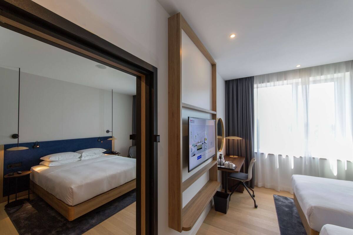 A hotel room with a large bed, a desk with a chair, a TV mounted on the wall, and a large window with curtains.