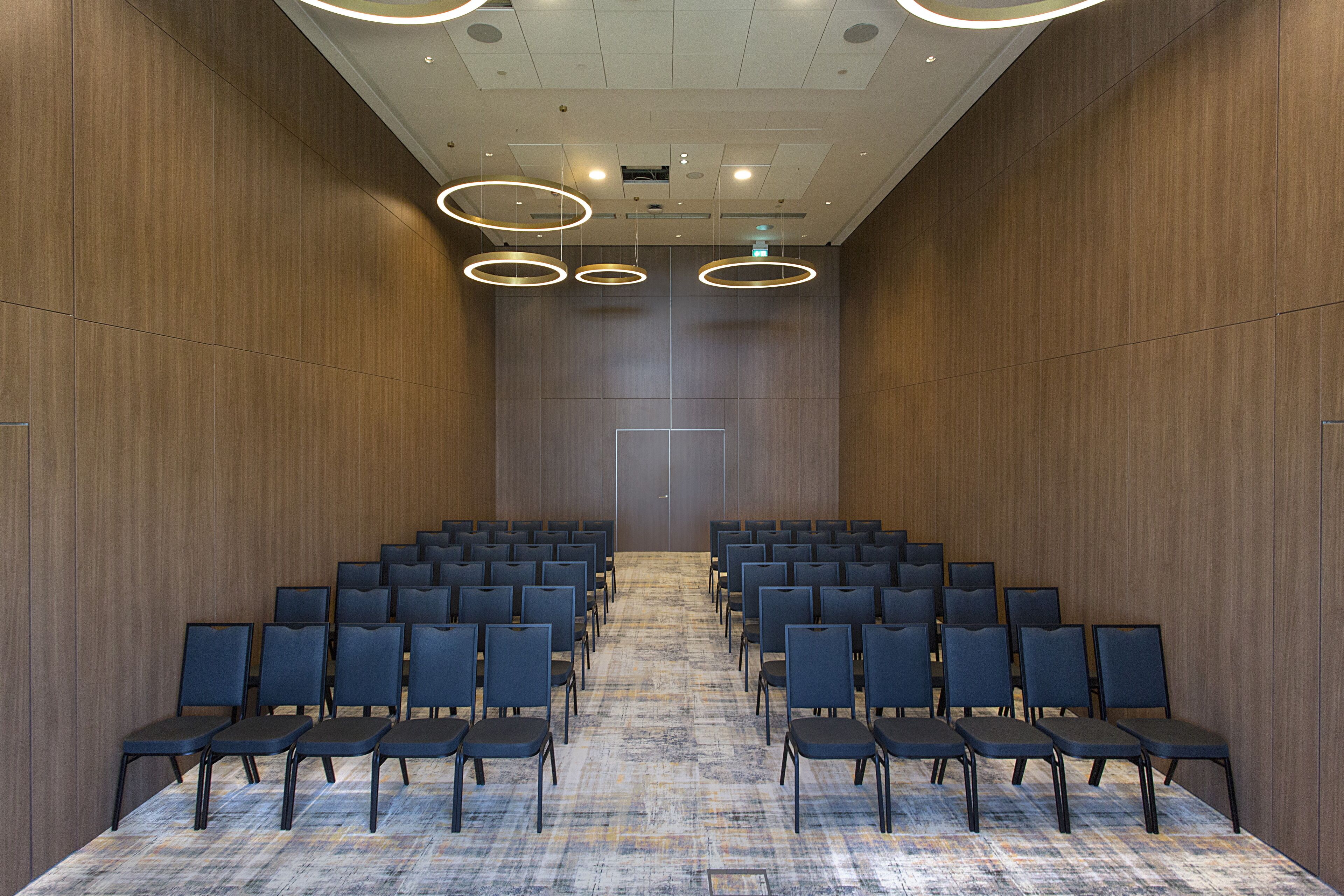 Ein Konferenzraum mit Reihen von Stühlen, einem gemusterten Teppich, Holzwänden und runden Deckenleuchten.