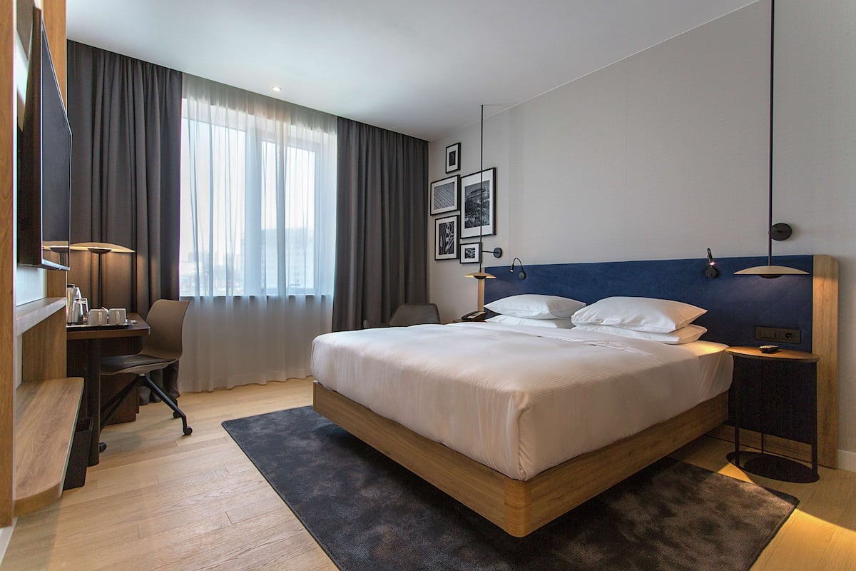 A modern hotel room with a large bed, a desk with a chair, a small table, and a window with curtains.