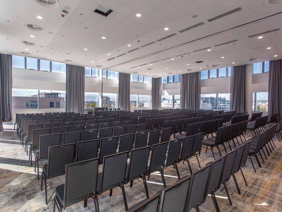 Een vergaderzaal met rijen stoelen, grote ramen en uitzicht op de stad.