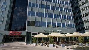 Ein modernes Hotel mit Glasfassade, einer Sitzgelegenheit im Freien inklusive Sonnenschirmen und einem gut sichtbaren Schild von Hilton Garden Inn.