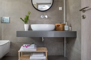 A modern bathroom with a large white sink, a round mirror, a potted plant, and a wooden shelf with towels. - E & G Relax Homes (Kissamos)