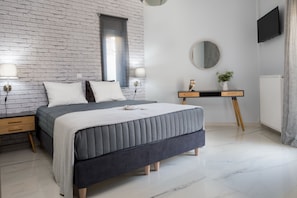 A modern bedroom with a brick wall, a large bed, bedside tables, a desk, a round mirror, and a TV. - E & G Relax Homes (Kissamos)