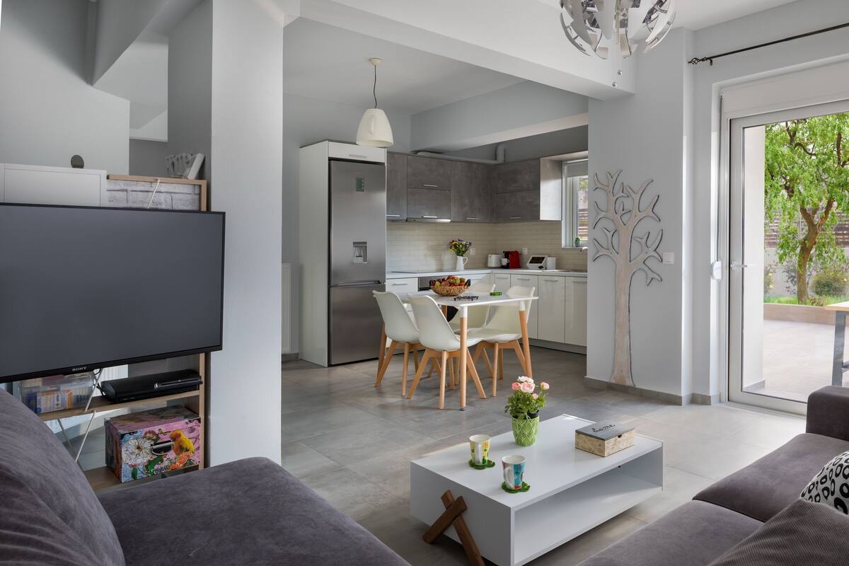 A modern living room with a flat-screen TV, a white coffee table, and a sofa. There is a dining area with a white table and chairs, and a kitchen with grey cabinets and a stainless steel refrigerator.