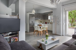 A modern living room with a flat-screen TV, a white coffee table, and a sofa. There is a dining area with a white table and chairs, and a kitchen with grey cabinets and a stainless steel refrigerator. - E & G Relax Homes (Kissamos)