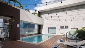 Un área de piscina en la azotea con terraza de madera, sillas de descanso y luces colgantes.