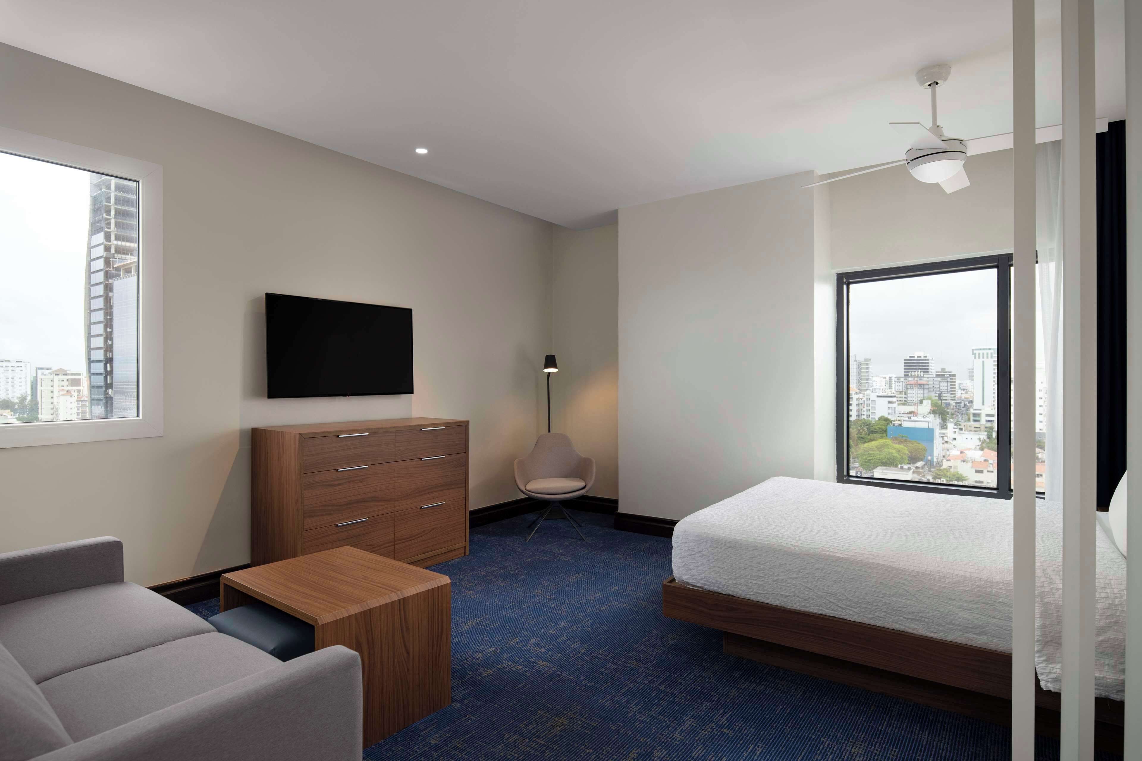 A modern hotel room with a bed, a desk, a chair, a dresser, a flat-screen TV, and a city view.