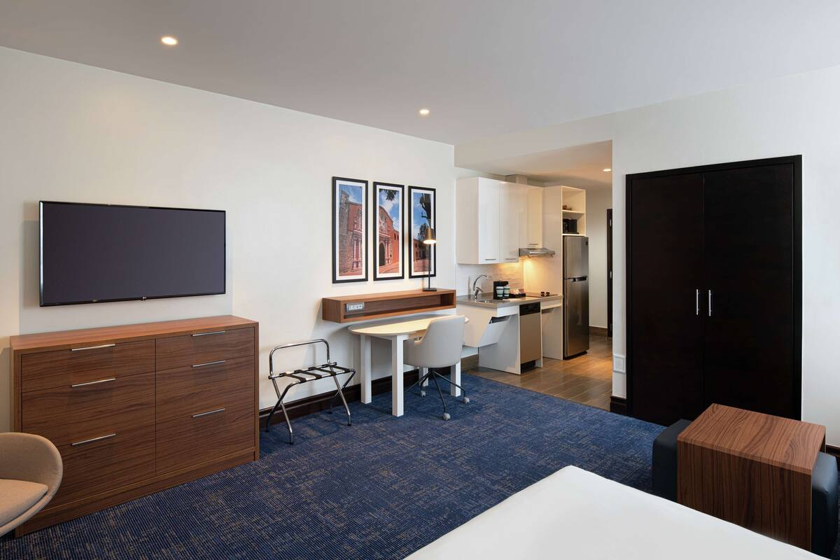 Ein modernes Hotelzimmer mit Flachbildfernseher, Schreibtisch, Stuhl, Kleiderschrank und einer Küchenzeile im Hintergrund.
