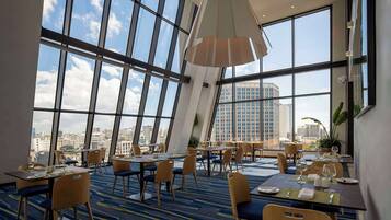 A modern hotel dining area with large windows offering city views, neatly arranged tables with chairs, and a blue carpeted floor.