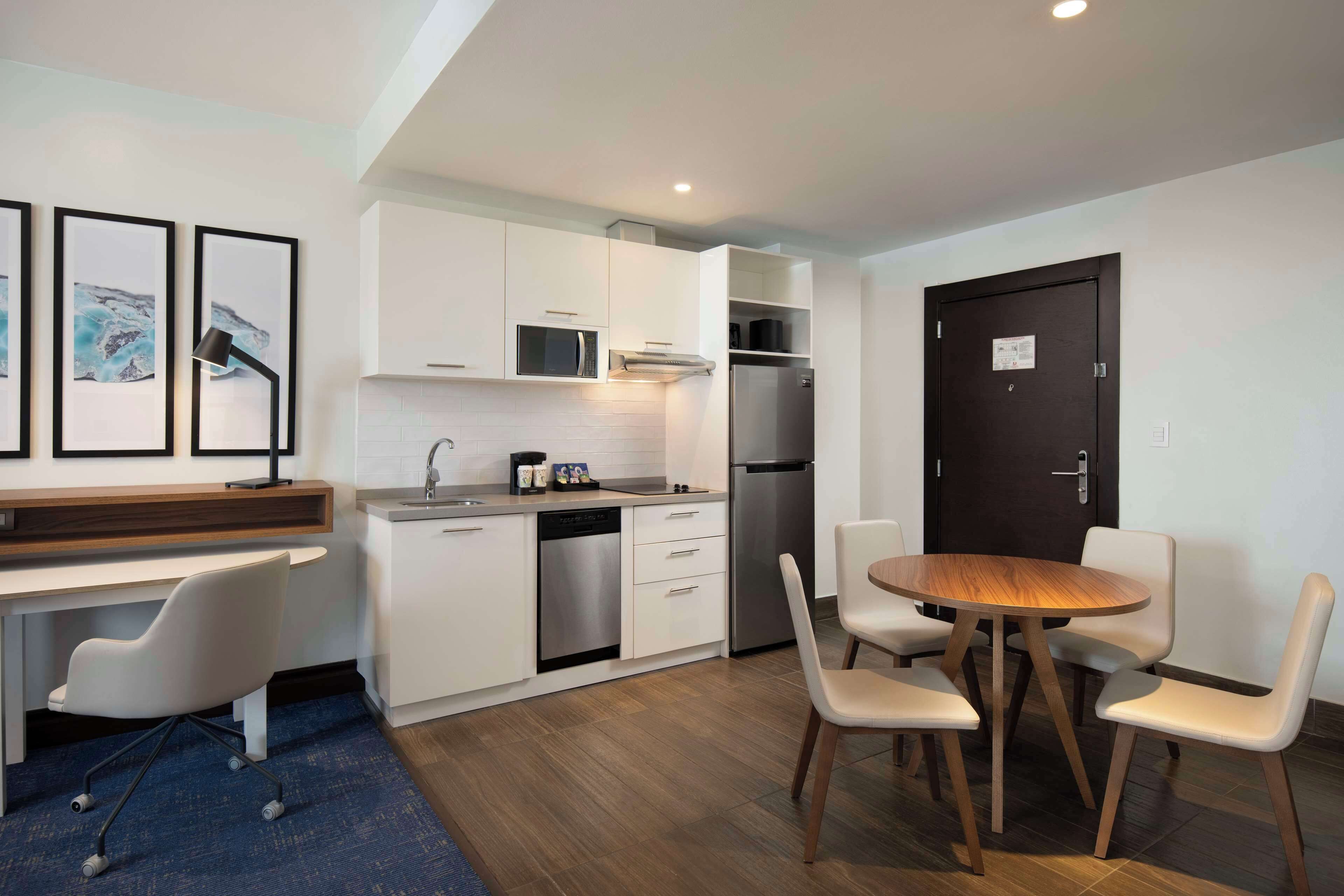 Una cocina moderna con armarios blancos, un refrigerador de acero inoxidable, un microondas y un lavavajillas. Hay una zona de comedor con una mesa redonda de madera y cuatro sillas.