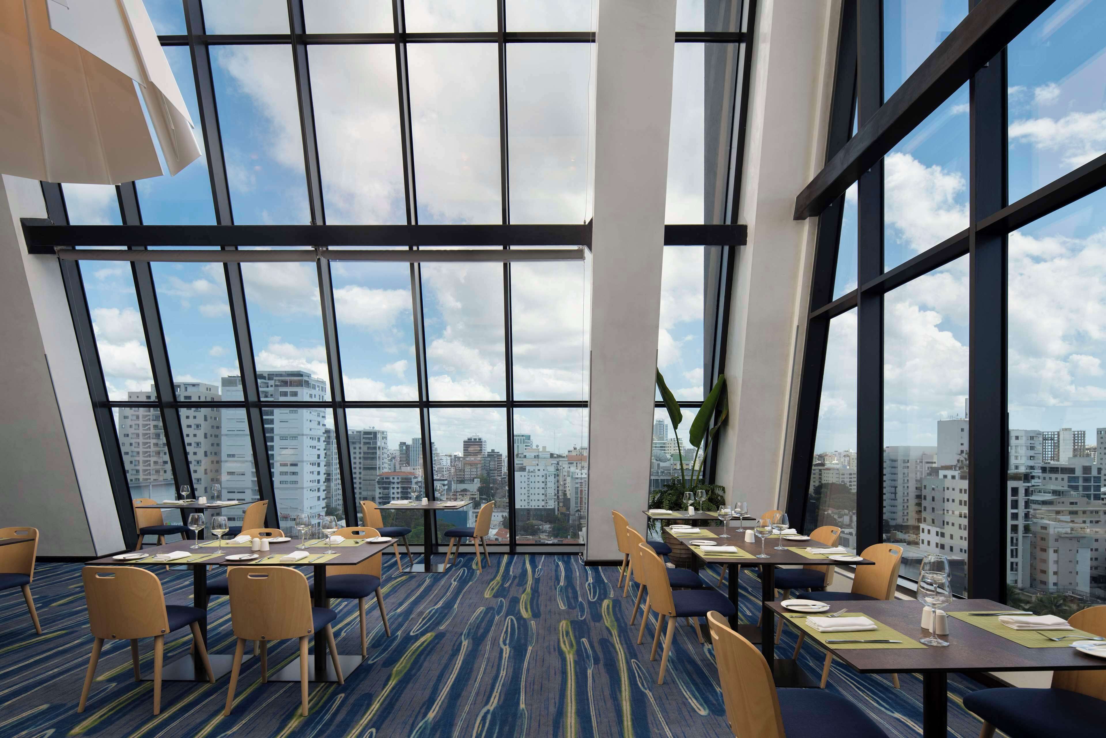 A modern hotel dining area with large windows, city views, and a blue carpeted floor.