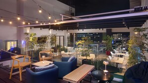 Un espacio al aire libre con mesas y sillas de madera, sofás azules y plantas en macetas.